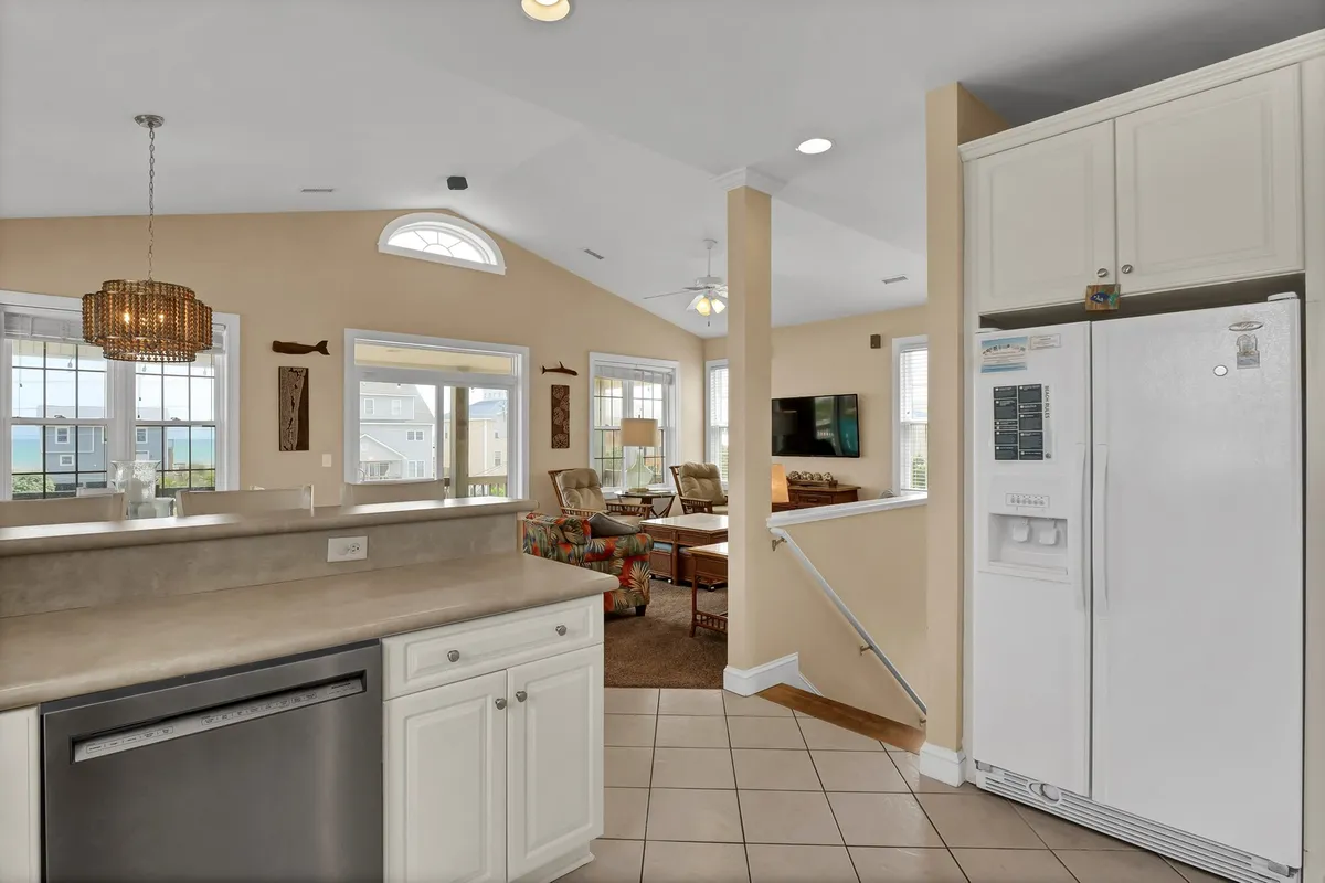 This image presents a bright, open-concept interior where the kitchen island flows into the living area, all bathed in natural light from large windows. The image showcases a cohesive space ideal for cooking, socializing, and relaxing, with both the kitchen and living room visible. The expansive windows hint at the scenic views beyond and reinforce the sense of gathering and openness described in the section.