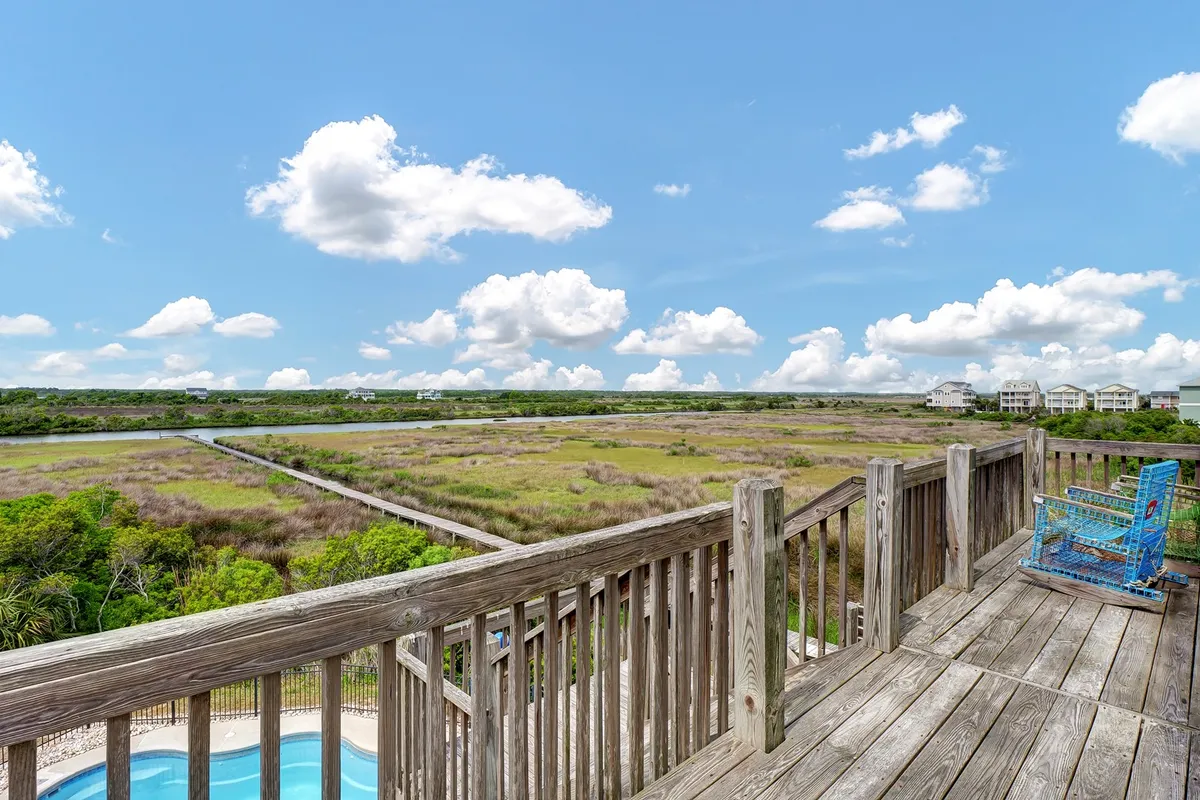 A tranquil marshland stretches beyond a wooden deck railing, with a colorful chair inviting visitors to relax and take in the peaceful scene. The expanse of blue sky and distant greenery hint at the quiet beauty of the sound side of the property.