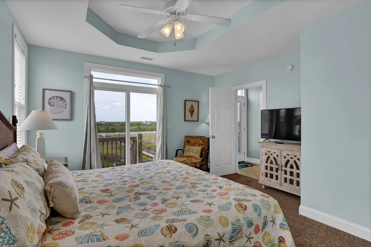 A cozy bedroom with a colorful quilt on the bed, a large window allowing natural light to fill the space, a comfortable chair, and a television. The window provides a scenic view, creating a restful environment.