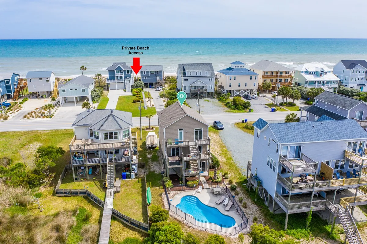 The image showcases the exterior of a beach house with a private beach access sign, a shimmering swimming pool in the foreground, neighboring houses, and sweeping ocean views in the background. The overall scene conveys a relaxed, inviting atmosphere perfect for outdoor enjoyment and seaside relaxation.