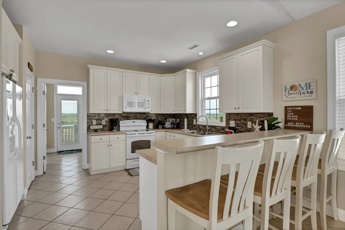 A homey kitchen scene centers on a kitchen island with stools, flanked by expansive cabinetry and large windows. The image suggests both functionality and casual connection, with opportunities for cooking, entertaining, and enjoying coffee with a view.