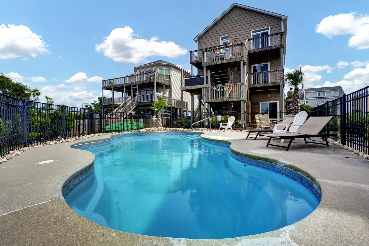 A sparkling swimming pool is surrounded by deck chairs and landscaping, positioned beside a multi-level house. The scene is bright and inviting, suggesting opportunities for group swimming, sunbathing, and al fresco meals. The setting is ideal for families or groups looking to enjoy shared poolside activities in a private setting.