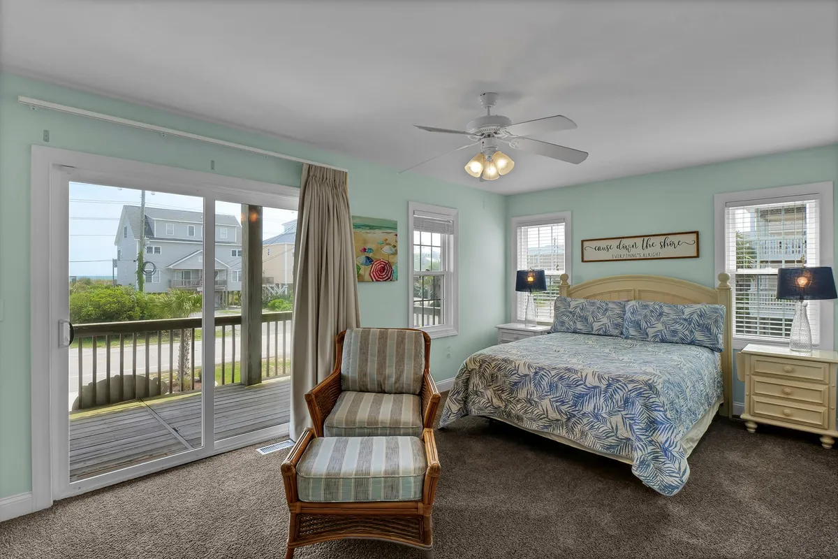 A tranquil bedroom featuring a bed with tropical-patterned bedding, sliding glass doors leading to a private deck, a comfortable chair, and decorative wall art. Sunlight streams in, highlighting ocean views just beyond the deck.