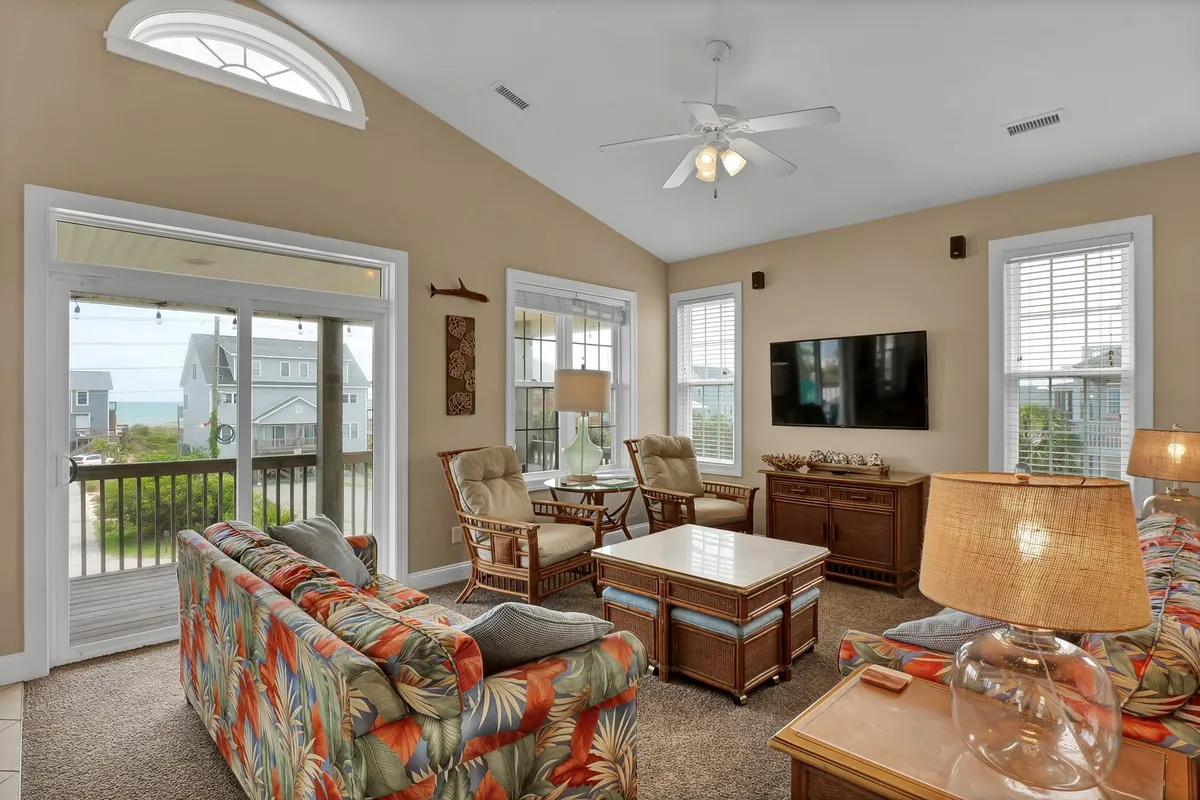 This living room photo features a colorful sofa, wooden coffee table, and large windows overlooking beautiful outdoor views. The television and ample seating suggest movie nights and group relaxation, while the expansive windows connect the space to the deck and beyond.