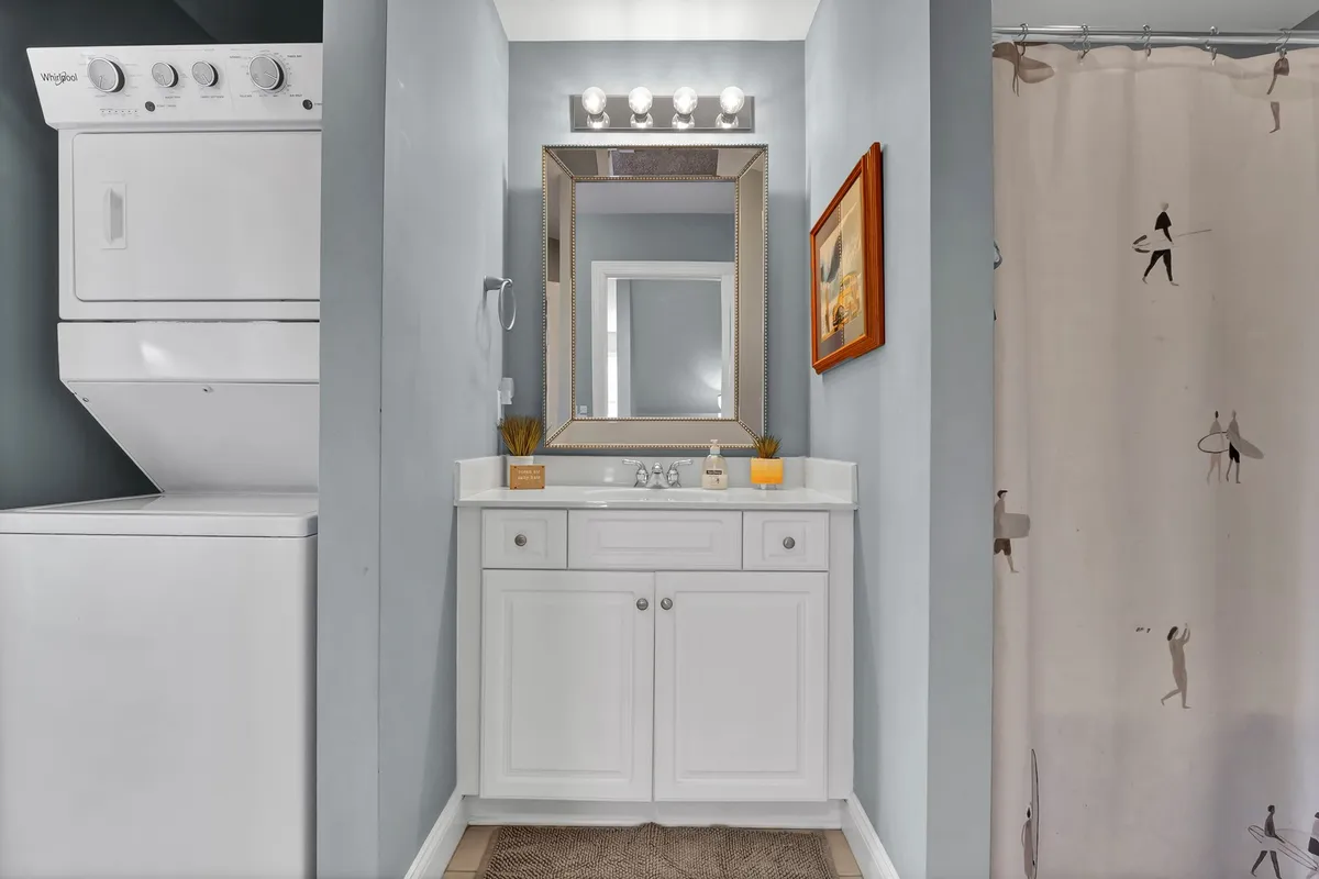 This image features a compact laundry area with a washer and dryer, vanity with mirror, decorative artwork, and a shower curtain. The space is practical, tidy, and includes storage options for linens and towels.